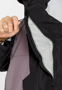 Zwarte en grijze zip-up jack, met een gevoerde binnenkant met geperforeerde textuur en een zijrits. De buitenstof lijkt waterafstotend te zijn.
