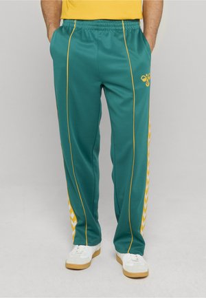 Türkisfarbene Jogginghose aus glattem Stoff, mit gelben Seitenstreifen und Akzenten, sowie einem Designlogo in Hüfthöhe. Mit elastischem Bund.
