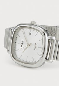 Montre Casio argentée avec un boîtier carré, un cadran blanc, des aiguilles argentées, une fenêtre de date et un bracelet en métal texturé. Résistante à l'eau jusqu'à 50m.