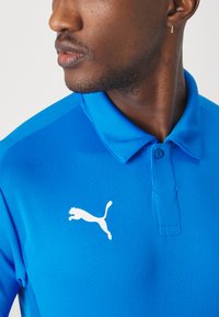 Modra polo majica iz teksturiranega blaga, z zavihanim ovratnikom in belim logotipom Puma na levem prsnem delu.