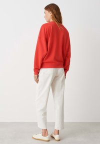 Sweatshirt rouge avec des épaules tombantes, jean blanc à coupe décontractée et baskets blanches avec une semelle beige. Tissu lisse et design simple.