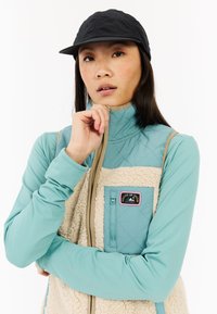 Lichtblauw zip-up vest met een gewatteerd ontwerp, lichtbeige fleece voorkant en zakdetails. Zwarte pet accentueert de outfit.