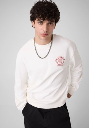 Jeune homme aux cheveux bouclés foncés porte un sweat-shirt blanc avec l’inscription rouge California Athletics Department et un pantalon noir, les mains dans les poches.