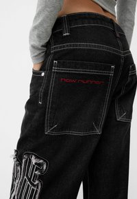 Mustat farkut valkoisella tikkauksella ja kulutetuilla yksityiskohdilla. Sisään ommeltu punaisella "new runner" -teksti taskussa.