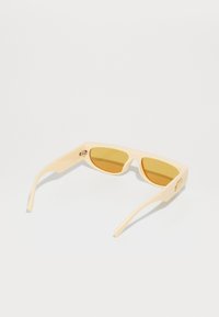 Occhiali da sole beige chiaro con lenti gialle, forma rettangolare, montatura in plastica liscia e aste sottili con piccoli dettagli di branding.