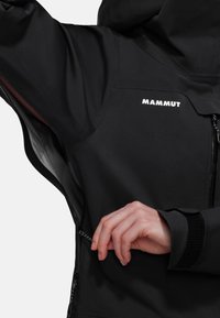 Svart vattentät jacka med en dragkedjeficka, elastiska ärmslut och "MAMMUT"-logotyp på övre bröstet. Slät textur, åtsittande design.