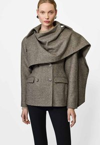 Dwurzędowy płaszcz wełniany w teksturowanym brązowym materiale, z opadającym kołnierzem i dużym krojem, z bocznymi kieszeniami i guzikami jako akcentami.