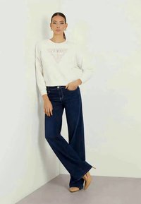 Ανοιχτού χρώματος φούτερ με γραφικό "GUESS", συνδυασμένο με σκούρες μπλε φαρδιές τζιν παντελόνες και γυμνά τακούνια, σε απλό φόντο.