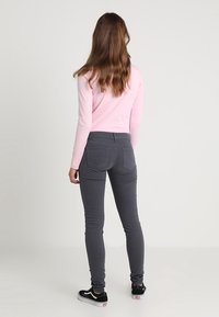 Dopasowane szare dżinsy z tylnymi kieszeniami, w połączeniu z długosłonym, jasnoróżowym topem. Modelka ma falujący włosy i nosi czarne trampki.