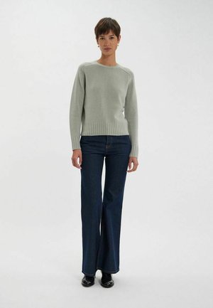 Pull en tricot gris clair avec un col rond et un ourlet côtelé, associé à un jean évasé bleu foncé et des chaussures noires.
