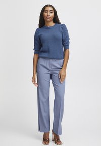 Blauer gestrickter Pullover mit strukturiertem Muster, kombiniert mit hellblauen maßgeschneiderten Hosen mit seitlichen Schlitzen und braunen hohen Schuhen.