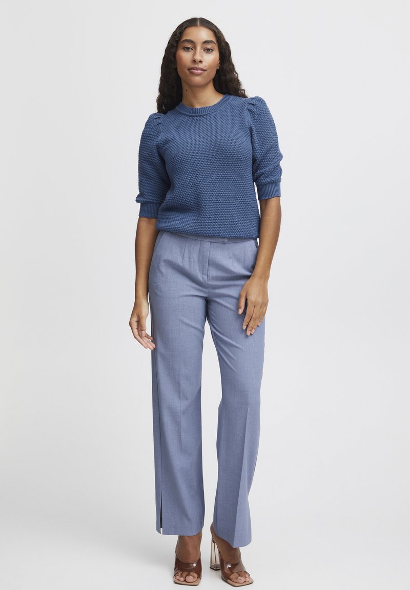 Blauer gestrickter Pullover mit strukturiertem Muster, kombiniert mit hellblauen maßgeschneiderten Hosen mit seitlichen Schlitzen und braunen hohen Schuhen.
