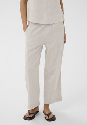 Personne portant un pantalon ample beige clair et un haut assorti, mains dans les poches, debout pieds nus dans des sandales noires à bout carré.