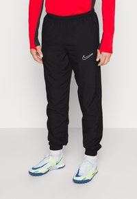 Zwarte sportieve broek met elastische tailleband, zijritszakken en een witte Nike-logo. Draagt bij kleurrijke, sportieve schoenen.