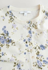 Cardigan floral en crème, orné de fleurs bleues et vertes. Texture douce avec un col et des poignets côtelés. Six boutons ronds sur le devant.