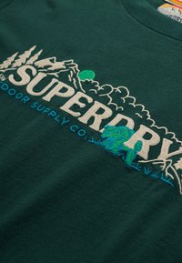 Zielona tkanina z haftowanym napisem "SUPERDRY", górami, drzewami, słońcem oraz niebieskim haftem "Outdoor Supply Co." poniżej.