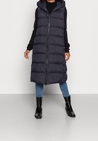 Lång, marinblå puffer-väst med huva, med hel frontdragkedja, vertikala quiltade sektioner och en något längre fåll. Bärs med svarta stövlar.