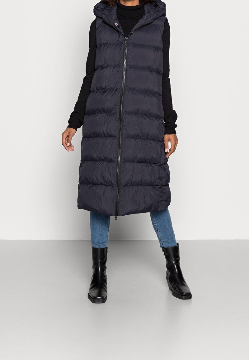 Lång, marinblå puffer-väst med huva, med hel frontdragkedja, vertikala quiltade sektioner och en något längre fåll. Bärs med svarta stövlar.