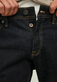 Jeans de ganga escura com costura amarela contrastante, fecho de botão e um pequeno logótipo metálico na cintura. Textura suave, corte justo.