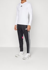 Witte lange mouwen sporttop met een V-hals en zwart Adidas-logo, gecombineerd met zwarte broek met witte strepen en een roze accent.