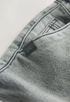 Material de blugi gri cu o textură netedă, având cusături contrastante și o etichetă mică neagră cu un detaliu în formă de stea.