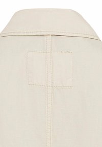 Beige skjorte med knapper og krave, med en central lodret søm og et firkantet lapdetalje på ryggen. Struktureret stof.