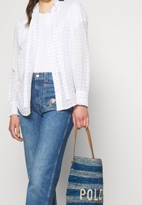 Blouse blanche à motifs avec des motifs bleus, associée à un jean bleu foncé avec broderies florales près de la poche, et un sac tissé bleu.