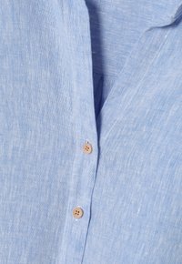 Camisa de lino azul claro con cuello, que presenta una tapeta de botones con botones color marrón claro, y un acabado de tejido texturizado.