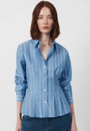 Kobieta ubrana w dopasowaną, niebieską koszulę na guziki z białymi pionowymi paskami i podwiniętymi rękawami, w połączeniu z niebieskimi dżinsami, stojąca na jednolitym tle.