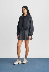 Μαύρο ελαφρύ μπουφάν bomber με ελαστικές μανσέτες, συνδυασμένο με γκρι πλισέ φούστα. Φορεμένο πάνω από μαύρη μπλούζα, ολοκληρωμένο με μπεζ αθλητικά παπούτσια.