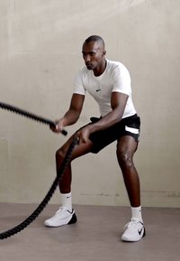 Mustat harjoitusnarut, joita pidetään henkilön toimesta, joka on pukeutunut tiukkaan valkoiseen Nike-paitaan ja mustiin shortseihin, valkoisten Nike-kensojen kanssa, tasaisella lattialla.