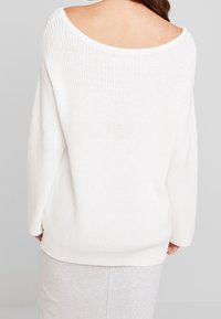 Žena v bílém pleteném svetru s širokým výstřihem a dlouhými rukávy, zobrazená zezadu na jednobarevném pozadí.