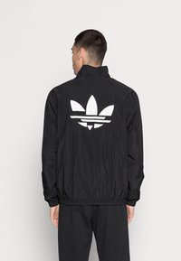 Svart lättviktsjacka med hög krage, elastiska muddar och en stor vit tre-strimlorslogo på baksidan. Slät textur.