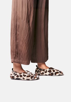 Pieds portant des chaussures à enfiler fourrées à motif léopard associées à un pantalon court marron ample sur un fond uni.
