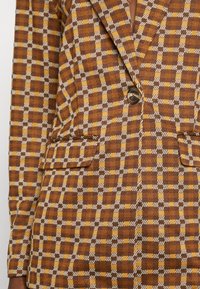 Bruine en gele geblokte blazer van textuurstof, met een notch-lapel en een enkele knoopsluiting. Voorzien van twee zakken aan de voorkant.