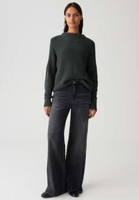 Donna che indossa un maglione lavorato a maglia verde scuro e jeans neri sbiaditi a gamba larga, in piedi contro uno sfondo semplice e chiaro.