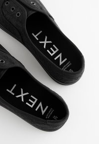 Sorte slip-on sko med mønstret stofoverdel, tekstureret overflade, øjeletsdetaljer og en kontrasterende sort indersål med sølvfarvet tekst.