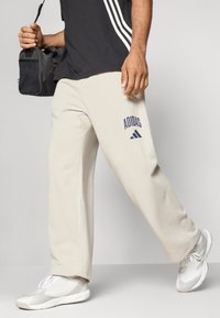 Homem a caminhar com calças de treino beige da Adidas, sapatilhas brancas, uma t-shirt preta com riscas brancas, e a carregar uma mala preta na mão esquerda.