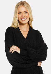 Schwarzer Strickpullover mit tiefem V-Ausschnitt, oversize Ärmeln und gerippten Bündchen. Texturiertes Zopf-Muster durchgehend. Sichtbare Accessoires und Schmuck.