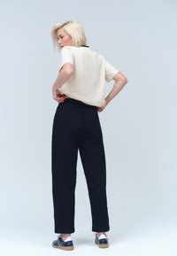 Beige lyhythihainen t-paita pyöreällä pääntiellä, yhdistetty tummansinisiin korkeavyötäröisiin housuihin. Malli käyttää tummia kenkiä ja on kuvattu selin kameraan.