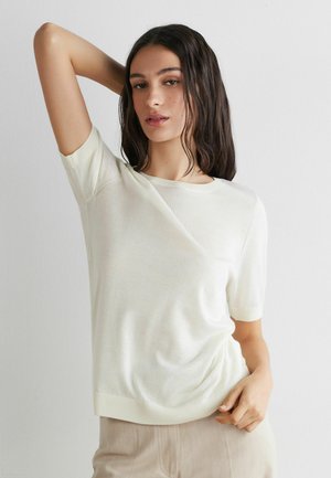 Femme aux longs cheveux foncés portant un haut en maille à manches courtes blanc et un pantalon beige, posant avec un bras levé derrière la tête.