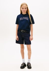 Μπλε ναυτικό βαμβακερό μπλουζάκι με λογότυπο «HILFIGER», συνδυασμένο με σκούρες τζιν σόρτς και μαύρα loafers με πλατφόρμα και λεπτομέρεια χρυσής αλυσίδας. Λευκά καλτσάκια αστραγάλου.