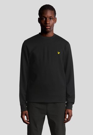 Man in een zwart shirt met lange mouwen en een klein geel vogellogo op de linkerkant van de borst, tegen een effen grijze achtergrond.