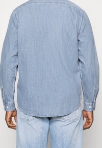 Světle modrá denimová košile s dlouhým rukávem, zahnutým lemem, zapínáním na knoflíky u manžet a mírně texturovanou látkou. Zobrazen pohled zezadu.