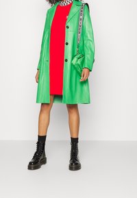 Zelený umělý kožený trenčkot s černými knoflíky, vrstvený přes červené šaty. Černé šněrovací kotníkové boty a malá zelená kabelka se strapcem.