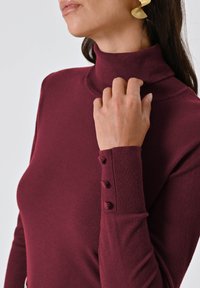Bordeaux coltrui met een slim-fit ontwerp, geribbelde manchetten en drie knoopaccenten op de mouw. Zachte textuur, lichtgewicht stof.