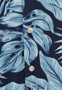 Korte mouwen shirt met een donker marineblauwe achtergrond, grote blauwe tropische bladeren en een witte knoop bij de opening.