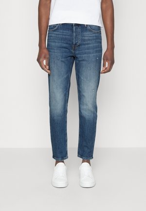 Mann in blauen Slim-Fit-Jeans mit leichtem Verblassen und weißen Sneakers, steht vor einem einfarbigen hellen Hintergrund.