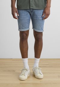 Denim shorts in light blue with rolled hems, worn with white crew socks and off-white sneakers. Wood floor and plain background.