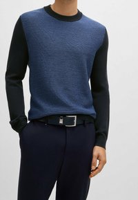 Marineblaues Sweatshirt mit einem strukturierten Fischgrätmuster an der Vorderseite und einfarbigen dunklen Ärmeln. Kombiniert mit dunklen Hosen und einem passenden Gürtel.